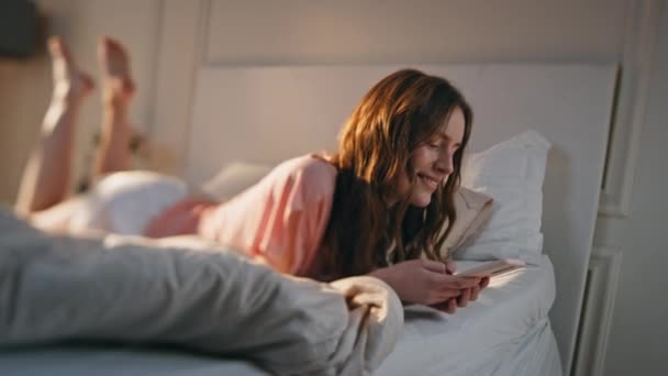 Smiling girl playing mobile phone games online at home. Relaxed teenager typing message browsing internet in morning sunlight. Carefree young woman lying bed look smartphone. Digital lifestyle concept - Footage, Video
