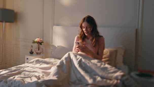 Wakker vrouw typt telefoon in de ochtend zonlicht. Glimlachend rustig meisje dat in bed ligt en online sms 't. Een ontspannen tiener die sms 'jes beantwoordt. Joyful vers vrouwelijk gebruik cel browsing web. - Video