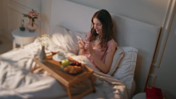 Tyyni nainen tekstailee kännykällä aamulla. Rento tyttö nauttia aamiaisen sängyssä selailu internetissä tarkistaa sosiaalisen median viikonloppuna. Kaunis rauhallinen nainen lähettää viestin älypuhelimella kotona. - Materiaali, video