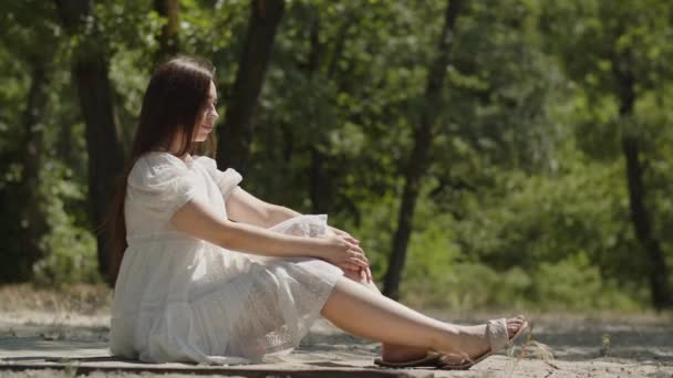 Красива дівчина в позитивних емоціях сидить збоку на піску в літньому лісі і медитує. Жінка в білій сукні посміхається, грайливо і кокетливо випрямляє волосся. Молода жінка позує - Кадри, відео