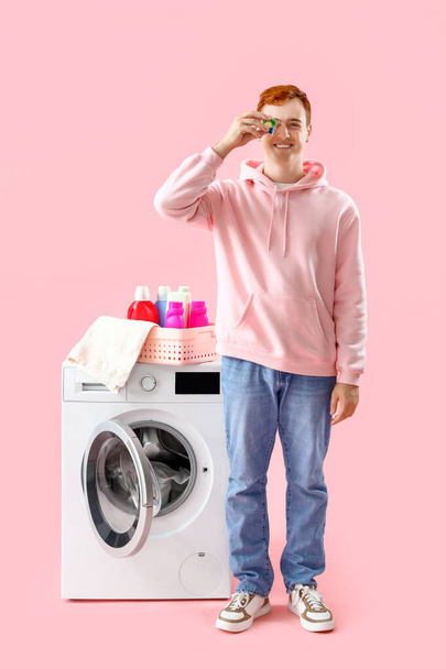 Hombre joven con cápsula de lavandería cerca de la lavadora sobre fondo rosa - Foto, Imagen