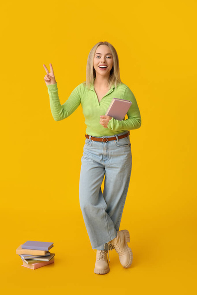 Beautiful young happy woman with many books showing victory gesture on yellow background - Photo, Image