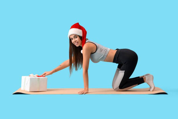 Sporty young woman in Santa hat with Christmas gift on blue background - Photo, Image