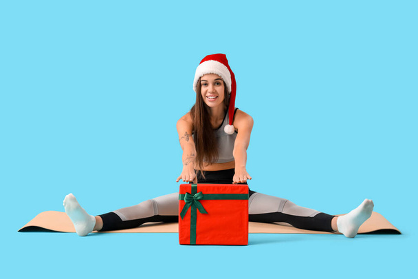 Sporty young woman in Santa hat with Christmas gift on blue background - Photo, Image