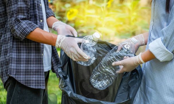 Рука тримає пластикові пляшки відходів, пара збирає сміття, прикладаючи до чорного сміттєвого мішка в парку соняшнику в день екологічної Землі. - Фото, зображення