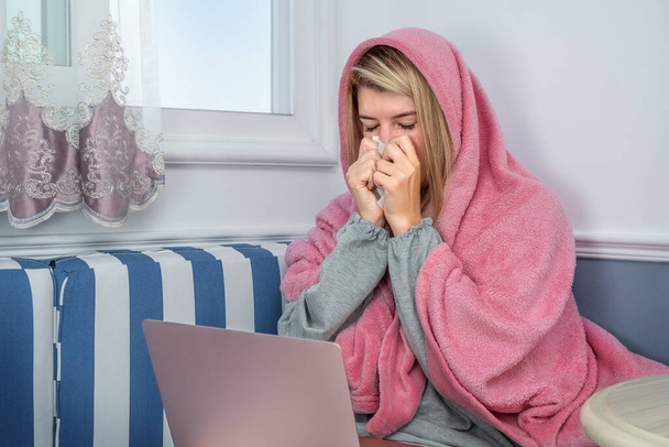 Uma menina espirra embrulhada em um cobertor e usa um laptop em casa - Foto, Imagem