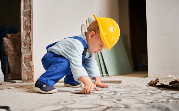 Trabalhador da construção menino instalando telha cerâmica no chão em apartamento em renovação. Criança usando capacete de segurança e macacão de trabalho enquanto joga em casa. - Foto, Imagem