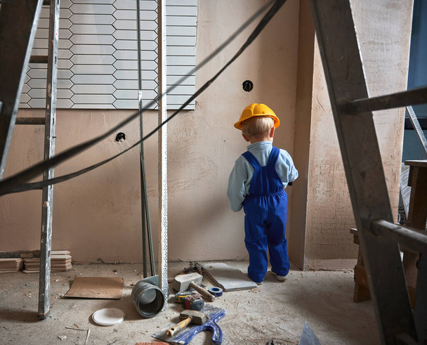 Dětský stavební dělník malování stěn s kartáčem v bytě při rekonstrukci. Zadní pohled na dítě nosí bezpečnostní přilbu a pracovní kombinézy při hraní doma. - Fotografie, Obrázek