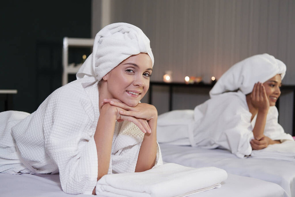 Uśmiechnięta biała i azjatycka kobieta w białym szlafroku i chustce na głowie relaksuje się razem na łóżku w spa. Salon piękności i styl życia lesbijek Concept. - Zdjęcie, obraz
