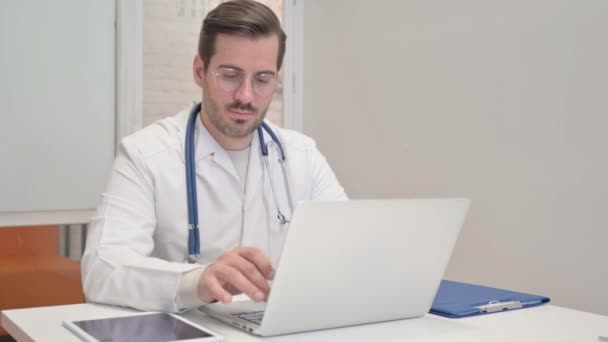 Médico de mediana edad mirando a la cámara mientras trabaja en el ordenador portátil - Metraje, vídeo