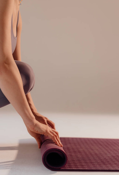 Una mujer joven gira una estera antes o después de una clase de yoga en el estudio. Piernas de cerca de una mujer haciendo yoga mientras está sentada sobre un fondo claro. - Foto, imagen