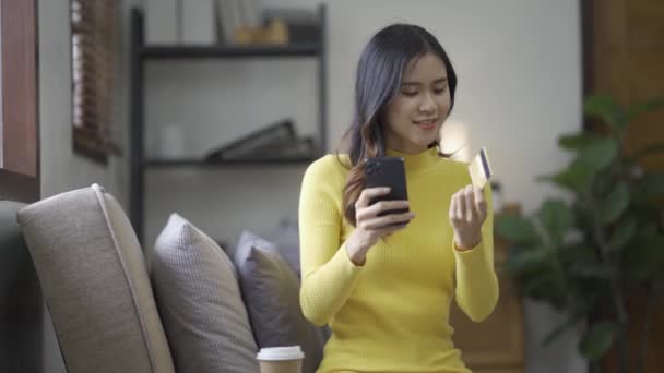 Kanepede otururken, elinde online alışveriş için kredi kartı tutan Asyalı bir kadın akıllı telefon kullanıyor ve evde internet üzerinden alışveriş yapıyor. - Video, Çekim