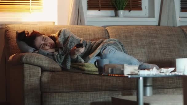 Проблема грипу та психології. Самотня молода кавказька жінка лежить на дивані вкритому ковдрою і перемикає телевізійні капелюхи з пультом дистанційного керування. Домашній вечір. Спостережувально-компульсивний розлад і депресія. - Кадри, відео