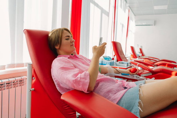 病院で献血をしている携帯電話を持った若い女性. 赤い心臓を抱えている間,女性が献血します. 献血について. - 写真・画像