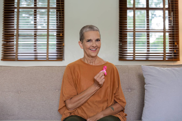 Portrait of confident smiling elderly senior woman with pink ribbon at her home. Health care, support, prevention. Breast cancer awareness month concept. - Photo, Image