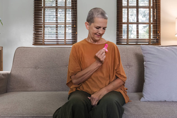Oudere vrouw met roze lint bij haar thuis. Gezondheidszorg, ondersteuning, preventie. Bewustmakingsmaand borstkanker. - Foto, afbeelding