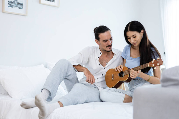 happy couple playing ukulele on bed in bedroom. man and woman enjoying playing music creates happy activities in family. young couple fun happiness romantic with instrument. - Photo, Image