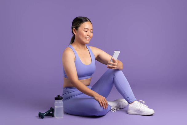 Mujer asiática en ropa deportiva se involucra con su teléfono celular, sentada y descansando después del entrenamiento en el suelo contra el fondo del estudio púrpura, ilustrando un descanso pausado - Foto, Imagen