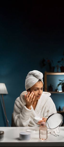 Mujer hispana en toalla de baño sonriente aplicando crema corporal facial en su cara para un rejuvenecimiento suave efecto hidratante. Banner vertical. Copiar espacio - Foto, Imagen