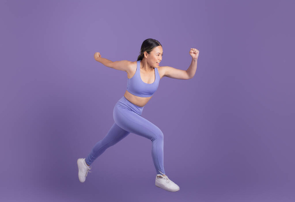 Joven mujer asiática en ropa deportiva corriendo en estudio sobre fondo púrpura, presentando imagen atlética clara y básica, tiro de cuerpo entero, espacio libre - Foto, Imagen