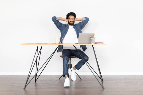 Un professionnel indien du millénaire, affichant un comportement détendu, apprécie son travail à distance, assis à une table confortable devant un ordinateur portable, incarnant un concept travail-vie équilibrée, appréciant les résultats - Photo, image