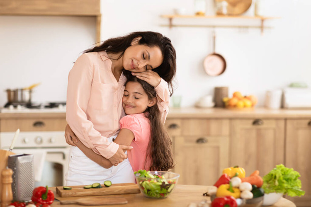 Mom and daughter kid sharing a warm embrace while cooking vegetable salad in modern kitchen at home, displaying affection and care amidst dinner preparation leisure - Photo, Image