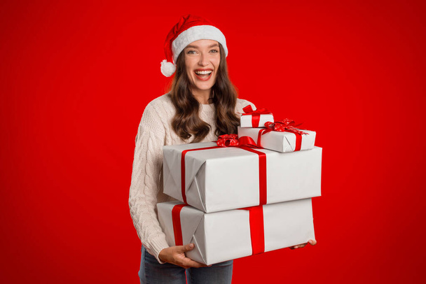 Радостная юная леди в шляпе Санта-Клауса, держащая две коробки с подарками на фоне красной студии, выражает позитивность и весёлые праздничные вибрации. Доставка и празднование рождественских подарков - Фото, изображение