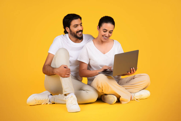 Famille adulte arabe heureux en t-shirt blanc assis sur le sol, utiliser un ordinateur portable, dactylographier, isolé sur fond de studio jaune. Appel vidéo, réunion, vente, achats en ligne et travail à distance ensemble - Photo, image