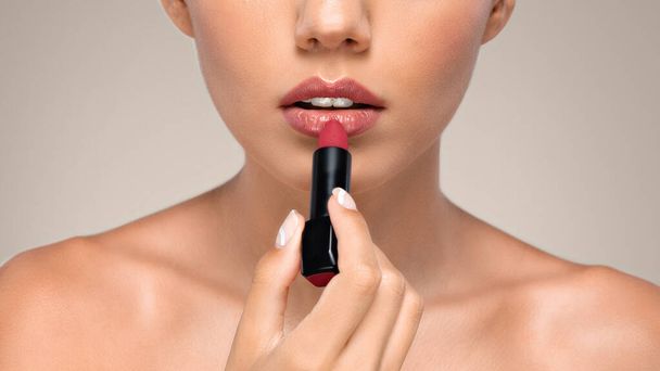 Kalme duizendjarige blanke vrouw met een perfecte huid aan te brengen rode lippenstift op de lippen, genieten van schoonheid les, geïsoleerd op grijze studio achtergrond, close-up. Basis naakte make-up, stijl en mode - Foto, afbeelding