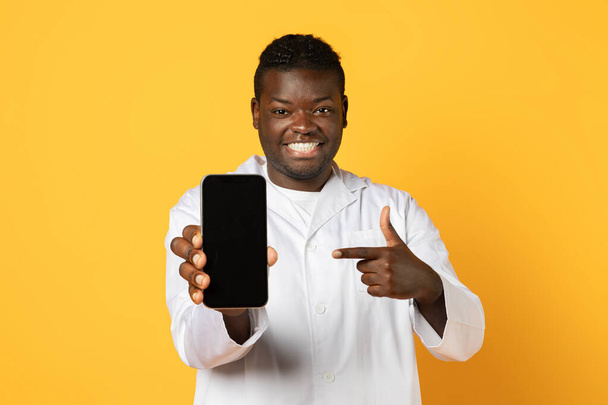 Медицинское приложение. Улыбающийся афроамериканец врач в униформе демонстрирует холостой смартфон перед камерой, доктор показывает пустой экран сотового телефона в руке над желтым фоном, макет - Фото, изображение
