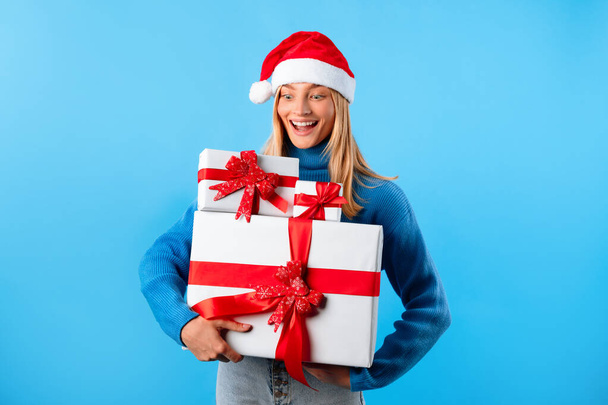 Noel Baba şapkalı heyecanlı kadın elinde bir sürü hediye kutusu, mavi stüdyo arka planında duruyor. Kış tatili satışları ve xmas teslimatı - Fotoğraf, Görsel