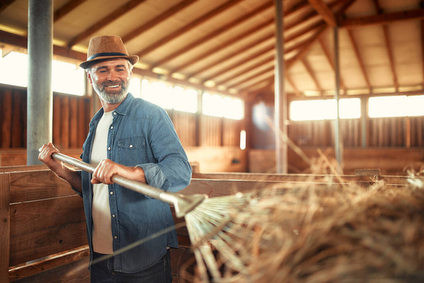 Mature handsome male farmer in a hat picking up hay with a pitchfork to feed livestock in a barn on a farm. Farming and agriculture concept. - Photo, Image