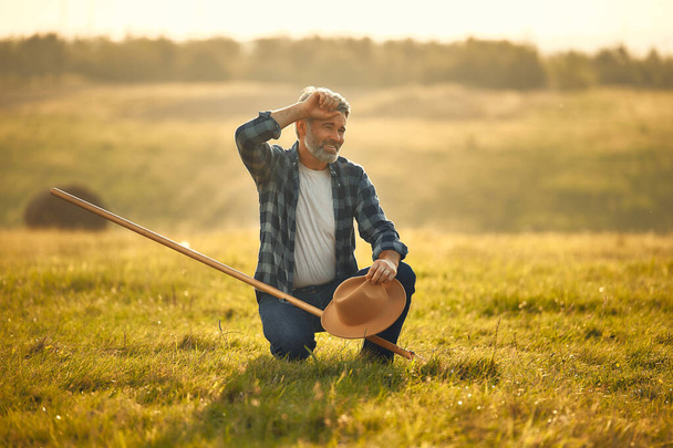 Volwassen knappe mannelijke boer met een hooivork tegen de achtergrond van een veld op een warme dag werken op een boerderij. Landbouw- en landbouwconcept. - Foto, afbeelding