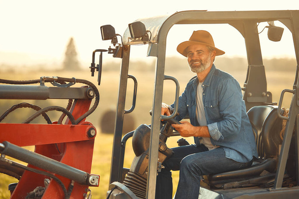 Volwassen knappe mannelijke boer zit in een tractor op een veld op een warme dag werken op een boerderij. Landbouw- en landbouwconcept. - Foto, afbeelding