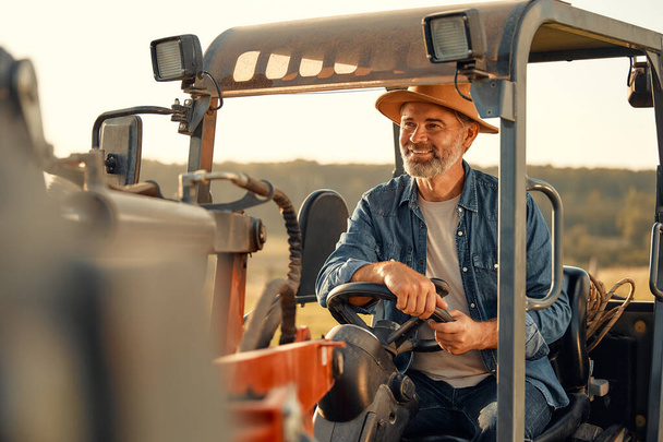 Olgun bir erkek çiftçi sıcak bir günde bir tarlada bir traktörde oturup çiftlikte çalışıyor. Çiftçilik ve tarım kavramı. - Fotoğraf, Görsel