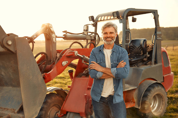 Зрелый красивый фермер, стоящий рядом с трактором на поле в жаркий день и работающий на ферме. Концепция сельского хозяйства. - Фото, изображение