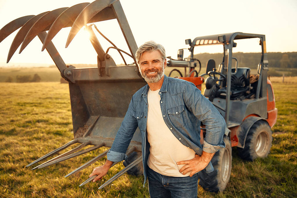 Kypsä komea miesmaanviljelijä seisoo traktorin lähellä pellolla kuumana päivänä ja työskentelee maatilalla. Maatalous ja maatalous. - Valokuva, kuva