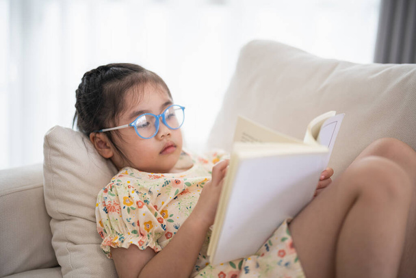 Radosny szczęśliwy azjatyckie dziecko dziewczynka uśmiecha się i czyta książkę, siedząc na kanapie w salonie w domu. Dziewczyny czytają książki uśmiech na kanapie w domu. Powrót do koncepcji szkoły. - Zdjęcie, obraz