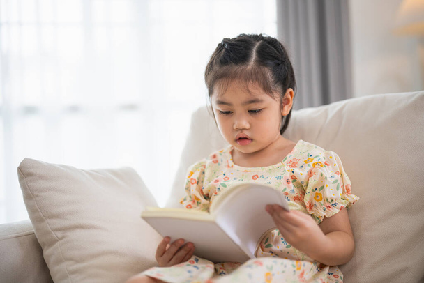 Vreugdevol gelukkig Aziatisch kind baby meisje glimlachen en lezen boek terwijl zitten op de bank bank in de woonkamer thuis. Een relex die een boek leest glimlacht op de bank in huis. Terug naar schoolconcept. - Foto, afbeelding