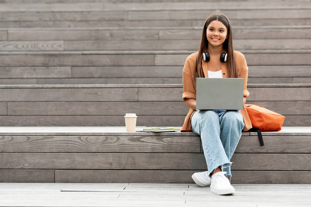 Jeune étudiante énergique assise seule dans les escaliers du campus universitaire vacant, s'engage avec son ordinateur portable, peut-être par appel vidéo ou assister à une conférence en ligne, faire des devoirs, copier de l'espace - Photo, image