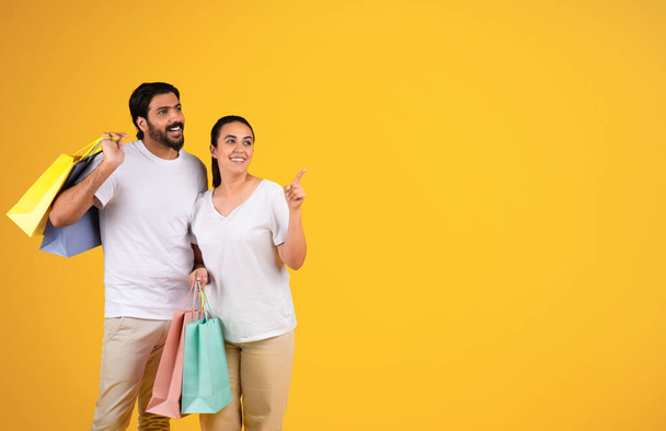 Lächelnder Millennial-Araber umarmt Ehefrau mit Tasche mit Einkäufen, zeigt mit dem Finger auf freien Raum, isoliert auf gelbem Studiohintergrund. Online-Einkaufsempfehlung, Bargeld zurück, Shopaholic in der Freizeit - Foto, Bild