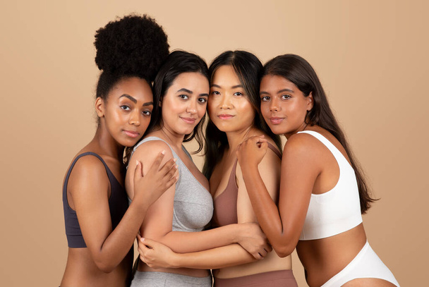 Κομψότητα προσωποποιημένη από τέσσερις διαφορετικές γυναίκες, αγκαλιάζοντας αρμονικά ενώ κοσμείται με απλά εσώρουχα, παρουσιάζοντας ταμπλώ φυσικής, ενιαίας ομορφιάς, μπεζ φόντο στούντιο - Φωτογραφία, εικόνα