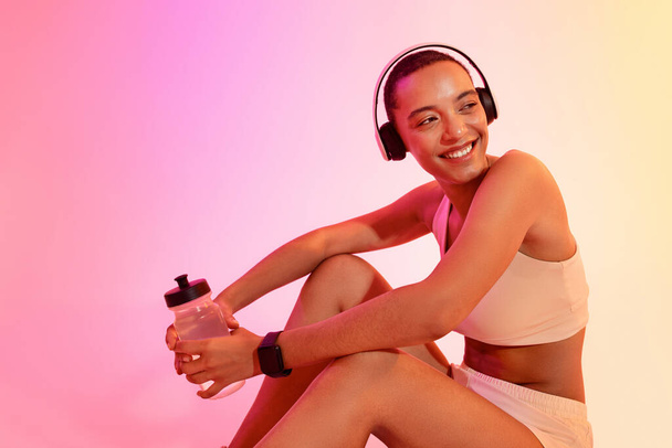 Χαρούμενη δραστήρια νεαρή Λατίνα γυναίκα σε αθλητικά ρούχα και ακουστικά με μπουκάλι νερό, σε νέον ροζ φόντο στούντιο. Γυμναστική με μουσική, αθλητισμό, φροντίδα του σώματος, απώλεια βάρους - Φωτογραφία, εικόνα