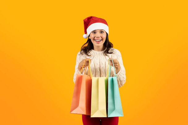 Xmas and winter holidays shopping. Positive young woman in Santa Claus hat holding Christmas gift bags over yellow studio background. New Year celebration and sales concept - Photo, Image
