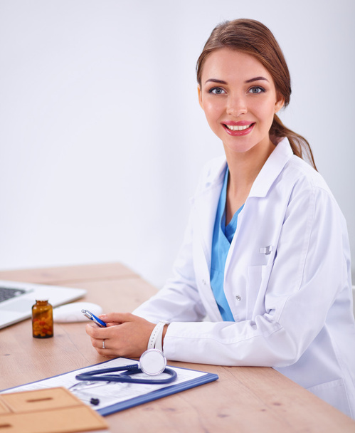 Beautiful young smiling female doctor sitting at the desk and writing. - 写真・画像