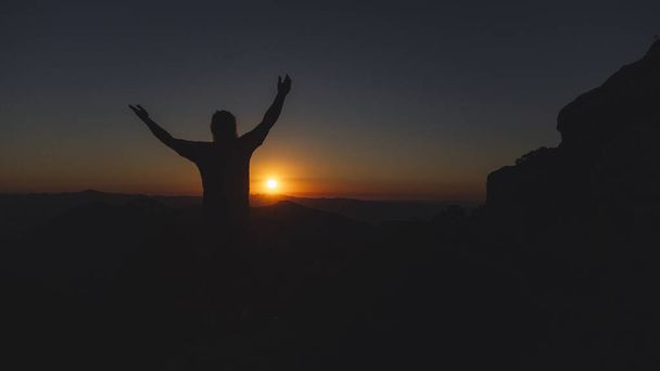 Ein Mann steht mit offenen Armen auf einem Berg, als die Sonne untergeht. - Foto, Bild