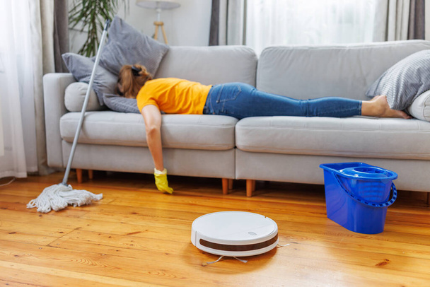 Robotyczny odkurzacz sprzątający pokój, podczas gdy zmęczona kobieta relaksuje się na kanapie po wykonaniu prac domowych. Koncepcja zalet nowoczesnych technologii czyszczenia, zakup odkurzacza robota - Zdjęcie, obraz