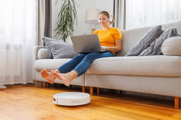 ロボット掃除機はリビングルームを掃除し,女性は自宅でソファに座ってラップトップを使用しています - 写真・画像