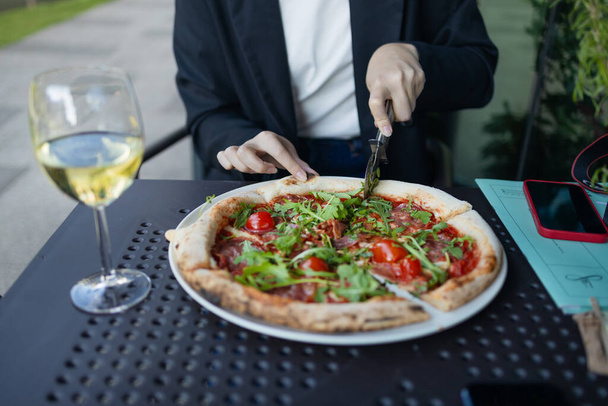 Detailní záběr kavkazské klientky ručně krájející pizzu s rajčaty, prosciuttem a rukolou s kulatým nožem na talíři v restauraci nebo kavárně. Italské rychlé občerstvení. Suché víno sklenice na - Fotografie, Obrázek