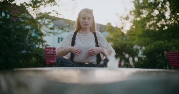 Giovane donna che medita mentre pratica yoga di fronte alla fontana al parco durante il tramonto - Filmati, video